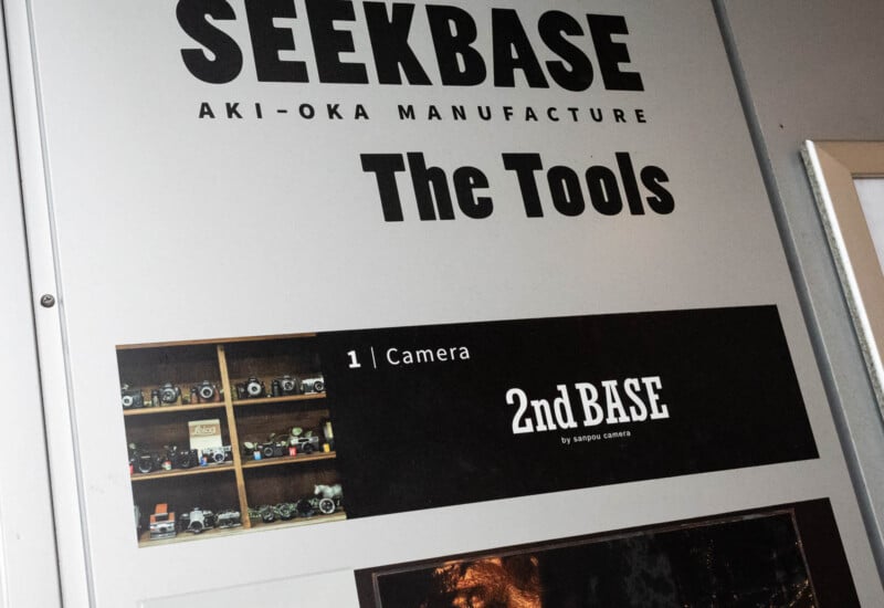 A signboard for SEEKBASE AKI-OKA MANUFACTURE featuring “The Tools” and “2nd BASE by sanpo camera,” with a photo of a shelf displaying various vintage cameras.