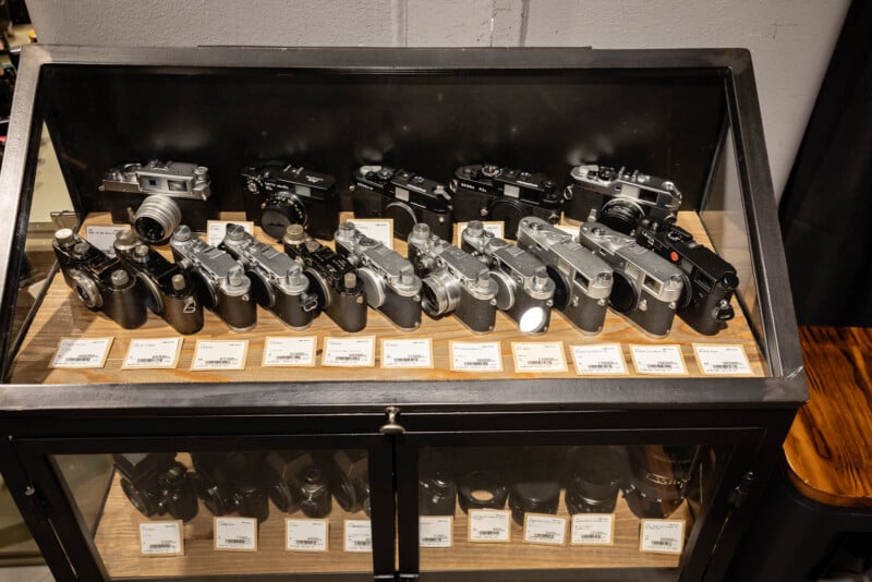 A glass display case containing two rows of vintage film cameras, each labeled with a small price tag. The cameras are mostly black and silver, and the case is set atop a wooden surface.