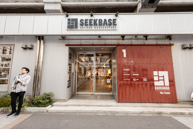 A man stands near the entrance of SEEKBASE, a modern building with a glass door and a large red sign displaying the SEEKBASE logo and tenant information. The building exterior is metallic and industrial in style.
