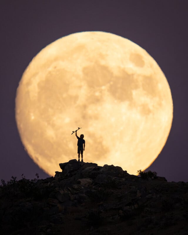 A person stands on a rocky hilltop, silhouetted against a large, bright full moon in the night sky, holding an object raised above their head.