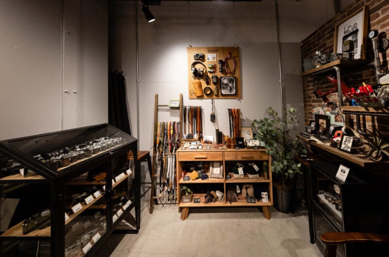 A well-lit shop interior with wooden shelves and a display case showcasing leather belts, watches, and accessories. Plants and decor items are arranged neatly on and around the shelving.