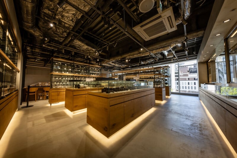 A modern, well-lit interior of a stylish store with wooden display cases, glass shelves holding various items, exposed ceiling ducts, and large windows letting in natural light.