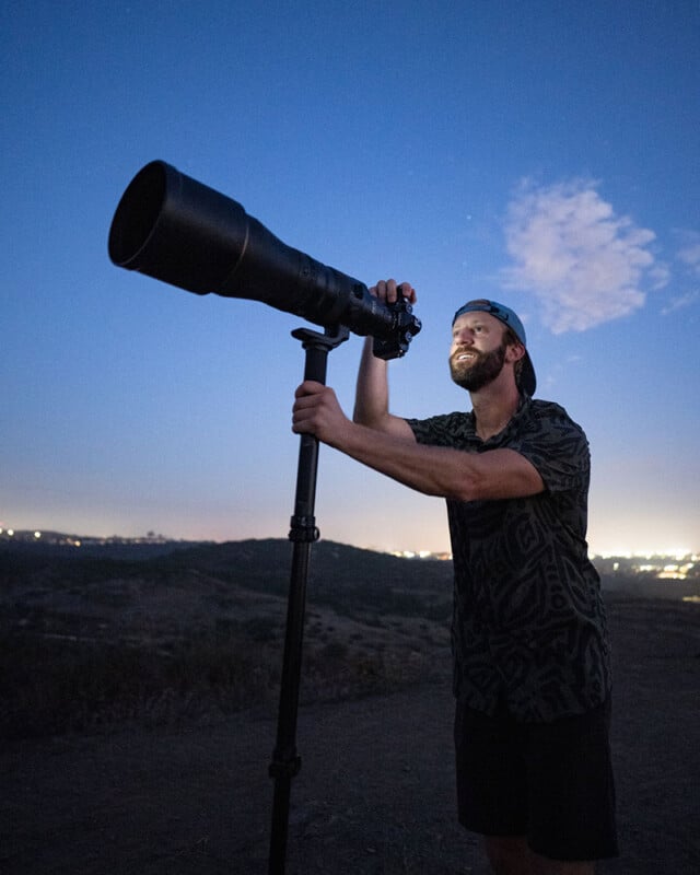 A man smiles while using a large camera with a telephoto lens mounted on a tripod, outdoors at dusk with city lights and a blue sky in the background.