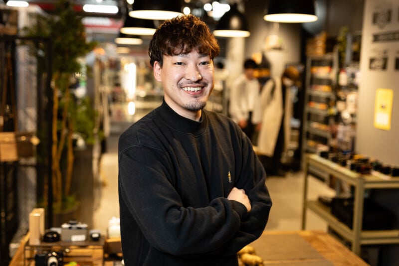 A man with curly hair and a beard smiles with arms crossed, standing in a well-lit store or workshop with shelves, hanging lights, and various items in the background.