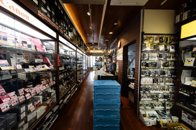 A narrow aisle in a camera store with shelves full of cameras and accessories. Two people stand at the counter near the center, and blue plastic crates are stacked in the middle of the wood-floored walkway.