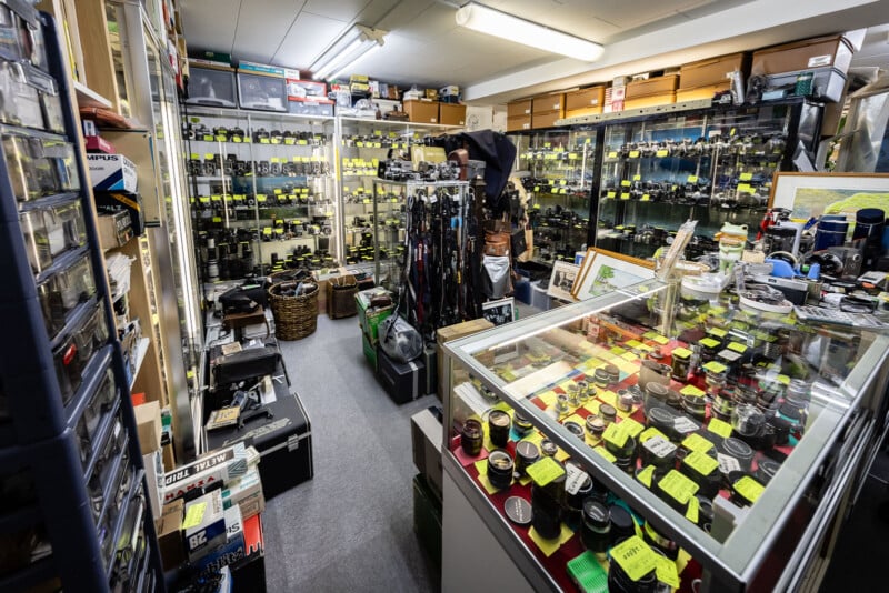 A cluttered camera and photography equipment shop with shelves and glass cases filled with cameras, lenses, and accessories, many labeled with bright yellow price tags. A person stands near the back of the store.