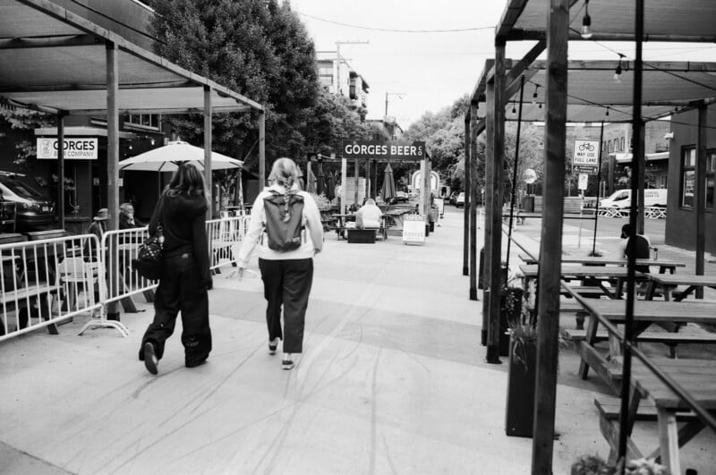 Two people walk down a city sidewalk beside outdoor seating areas with tables and benches. A sign reading "GORGES BEER" is visible ahead. Trees and buildings line the street in the background. The image is black and white.