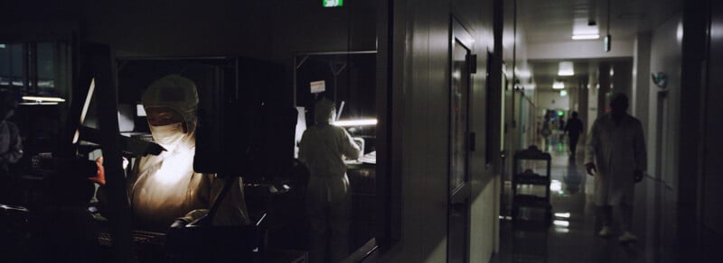 A person in protective lab gear works at a station in a dimly lit laboratory, while others in white coats and masks walk down a reflective hallway in the background.