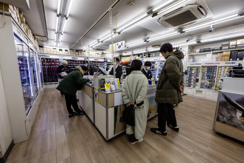 Several people stand at the counter inside a well-lit electronics or camera store, browsing products and interacting with staff. Shelves and display cases filled with equipment line the walls.