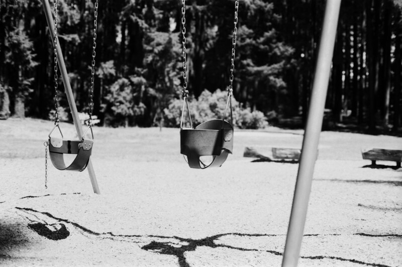Two empty toddler swings hang from chains at a playground. The area is covered in light gravel or sand with cracks on the ground. Trees and grassy areas are visible in the blurred background. The photo is in black and white.