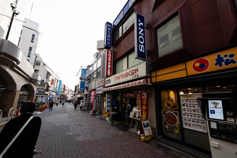 A street in Japan lined with shops and restaurants, including a Sony sign and a vending machine outside a restaurant. People are walking along the brick-paved sidewalk under a mostly cloudy sky.