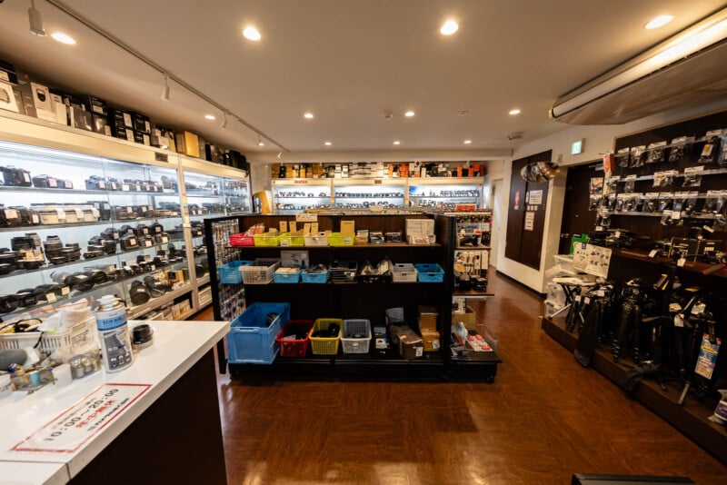 A well-lit camera store interior with shelves filled with cameras, lenses, and accessories. The center displays bins and boxes, while tripods and various equipment line the walls.