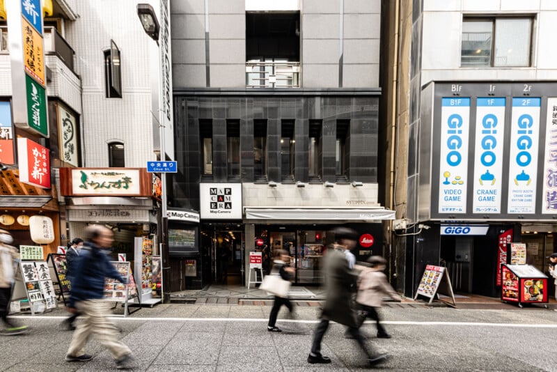 A busy street scene in Japan with blurred pedestrians walking past storefronts, including a café and arcades with blue "GIGO" signs, and various colorful shop signs on the buildings.