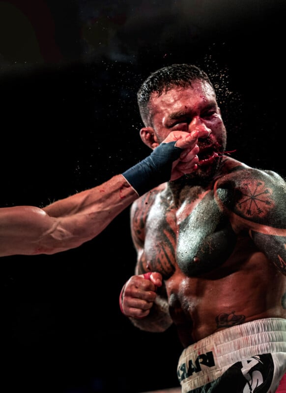 A boxer with tattoos gets punched in the face, sweat and possibly blood flying from his nose, during an intense boxing match under dramatic lighting.