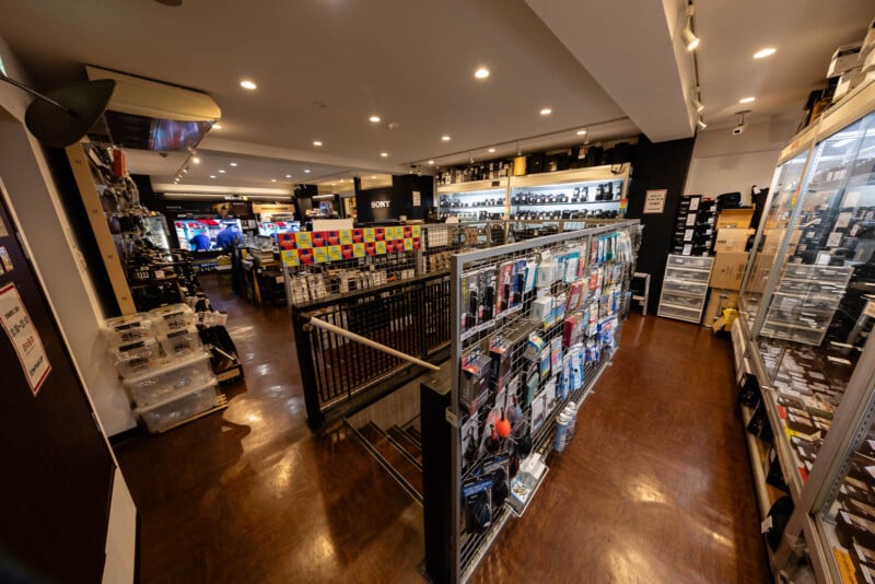 A well-lit camera store with shelves displaying various photography equipment, accessories, and cameras. The store has wooden flooring and multiple display cases organized neatly along the walls and aisles.