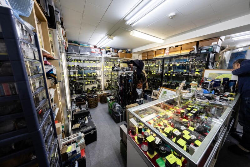 A small, crowded camera shop with glass display cases and shelves filled with cameras, lenses, and accessories. Yellow price tags are visible on many items, and two people are browsing the merchandise.