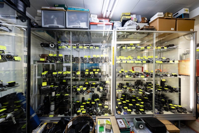 Glass display cabinets filled with vintage cameras, lenses, and accessories, each labeled with yellow tags. Shelves above and items below show more boxes, storage bins, and equipment in a cluttered room. Bright fluorescent lights above.