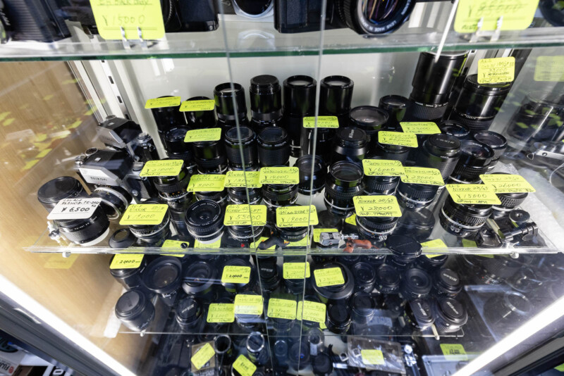 A glass display case filled with various camera lenses and a vintage camera, each item labeled with handwritten yellow price tags. The lenses are arranged on multiple shelves.