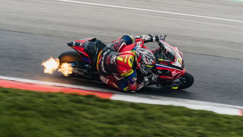 A motorcycle racer leans sharply into a turn on a track, with flames shooting from the exhaust. The rider wears a red and yellow suit and helmet, and the bike is red with sponsors’ logos. Grass and curbs line the track.