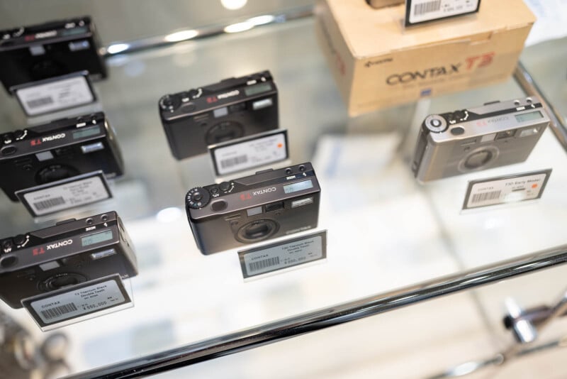 A glass display case showing several black film cameras, each labeled with price tags. A tan cardboard box labeled "Contax T3" is visible in the upper right corner of the image.