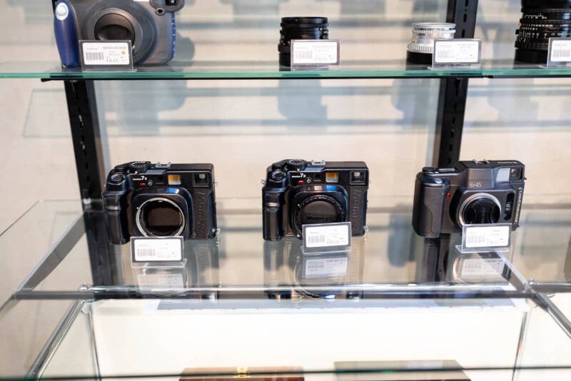 Three vintage medium format cameras are displayed on a glass shelf, each with a price tag. Additional camera equipment is seen on the shelf above, including camera lenses.