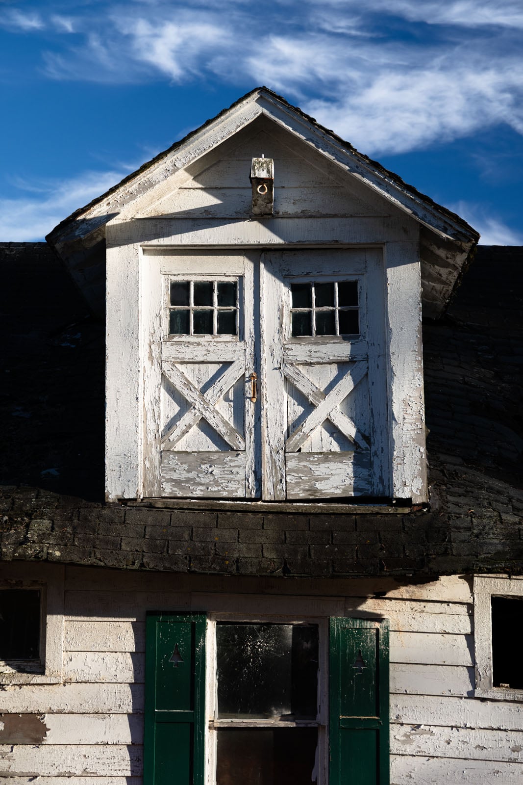 A close-up of an old, weathered white dormer with peeling paint and crossed wooden panels, sitting atop a similarly worn house against a blue sky with wispy clouds.