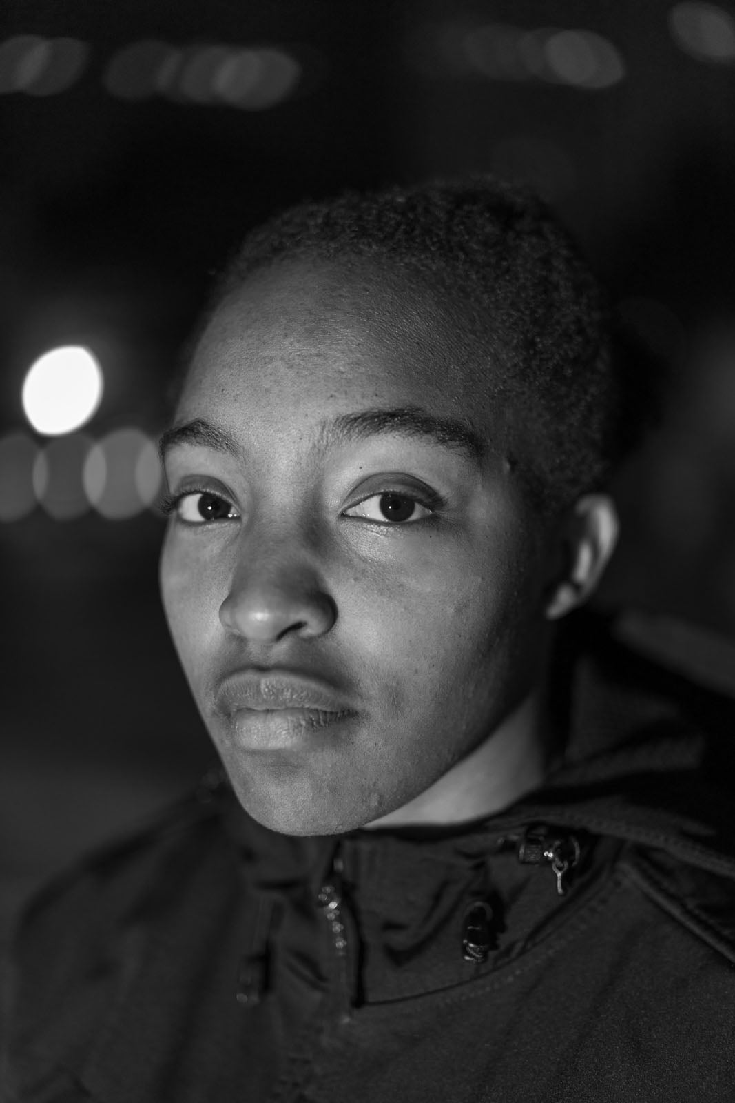 A person with short hair wearing a zip-up jacket looks directly at the camera with a neutral expression; the background is dark and blurred with visible lights.
