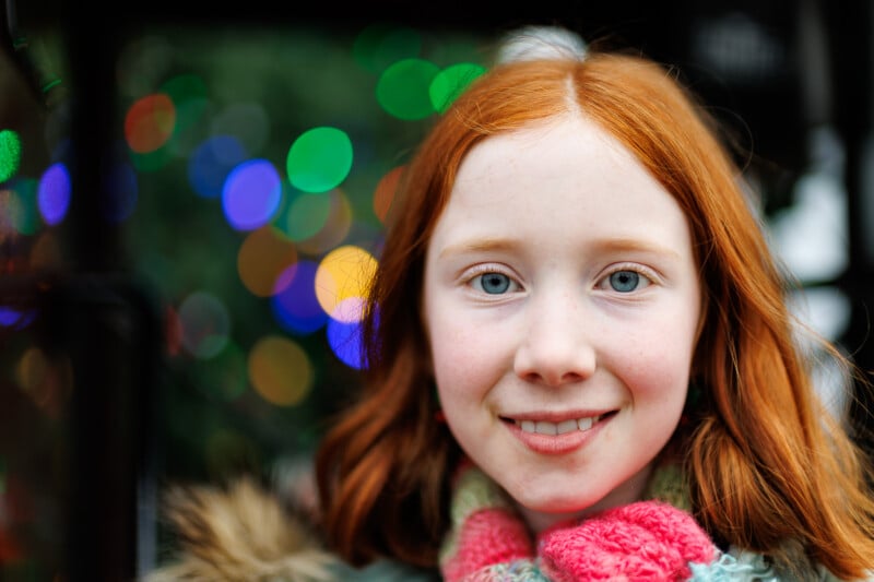 A young girl with red hair and blue eyes smiles at the camera, wearing a colorful scarf. Bright, blurred multicolored lights create a festive background.
