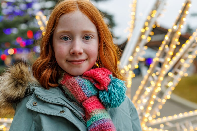 A young girl with red hair and a colorful scarf smiles in front of festive, glowing holiday lights outdoors. She is wearing a green coat with a fur-lined hood. The background is blurred with lights and trees.