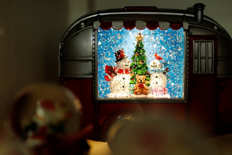 A festive snow globe scene inside a trailer, featuring two snowmen in hats and scarves, a Christmas tree, and a teddy bear, with snow falling around them and a blurred foreground.