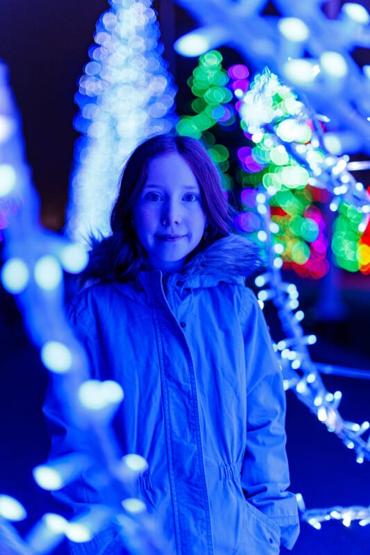 A child in a light blue winter coat stands among bright, colorful holiday lights with a blue and purple glow, surrounded by blurred decorations and illuminated trees in the background.