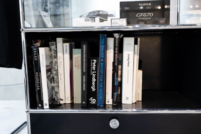 A black shelf contains neatly arranged photography and art books, with visible titles such as "Shoot for the Moon," "Peter Lindbergh," and "Exiles." A Fujifilm GF670 camera box sits on the top shelf.