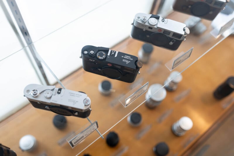 Several vintage Leica cameras are displayed on a glass shelf, viewed from above, with price tags beside them. Out-of-focus camera lenses and equipment are visible on the wooden surface below.
