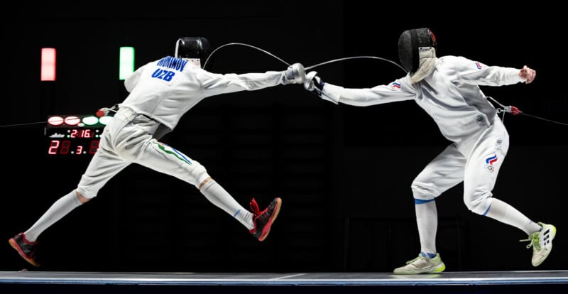 Two fencers in protective gear lunge towards each other, crossing swords mid-air on a fencing strip. Both are in dynamic poses, with one fencer’s foot off the ground, and a scoreboard visible in the background.