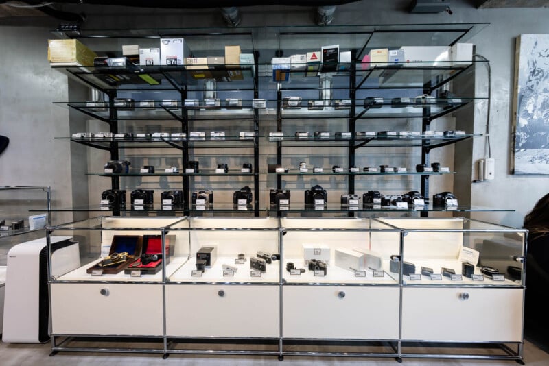 Glass display cases and shelves filled with various cameras, camera lenses, and accessories are arranged neatly in a modern store with a concrete wall background.