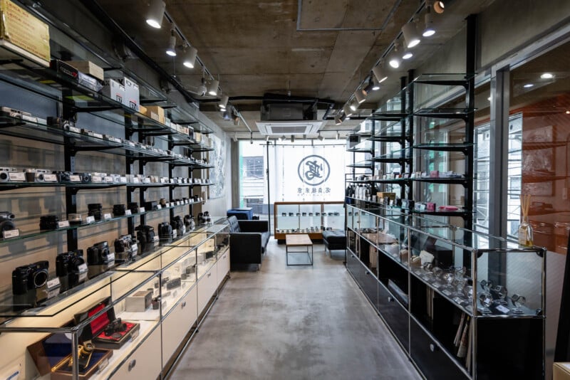 Modern camera shop interior with glass display cases and shelves filled with cameras and lenses, polished concrete floor, track lighting, and a large logo on the back wall near seating area.