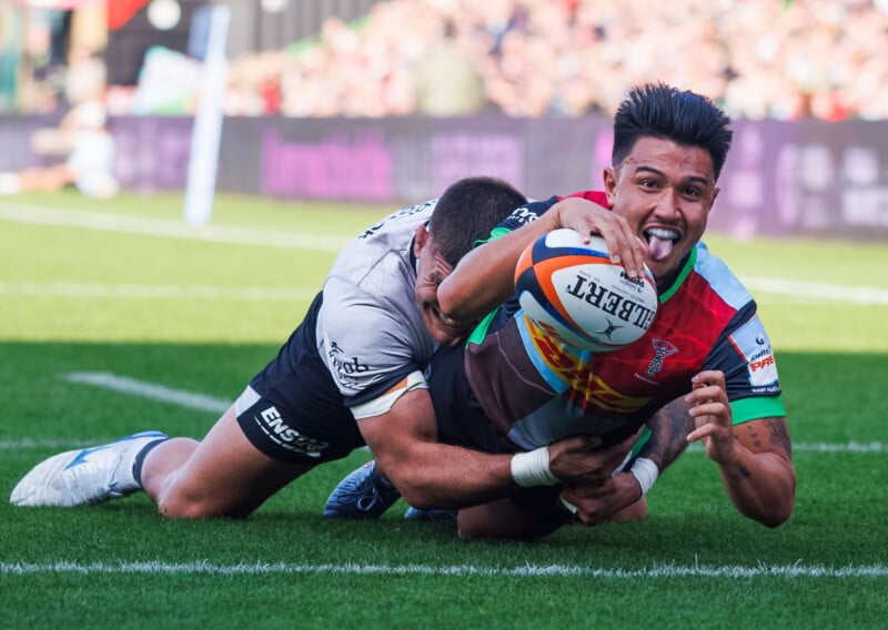 A rugby player in a colorful jersey stretches to score a try while being tackled by an opponent in a white jersey on a grassy field, with a crowd blurred in the background.