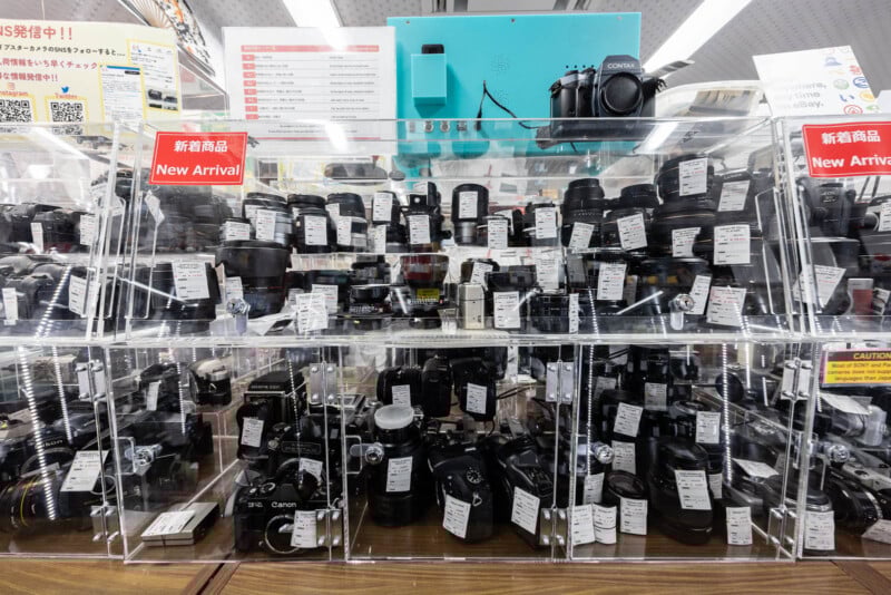 A display case in a store showcases various cameras and camera lenses, each with price tags attached. Signs reading “New Arrival” in English and Japanese are visible at the top of the case.