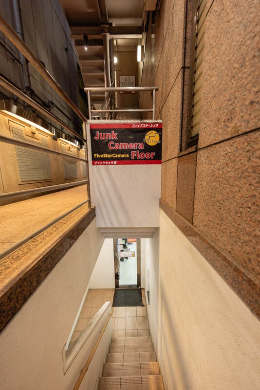 A stairwell in a building leads down to a door. Above the stairwell, a sign reads "Junk Camera Floor" and "FiveStarCamera" in English and Japanese. The walls and floor are tiled and clean.