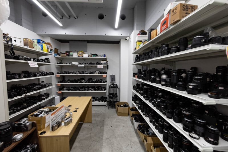 A room filled with shelves lined with various camera lenses and equipment, a wooden table in the center, and boxes on the floor; the space appears organized and well-lit with fluorescent lights.