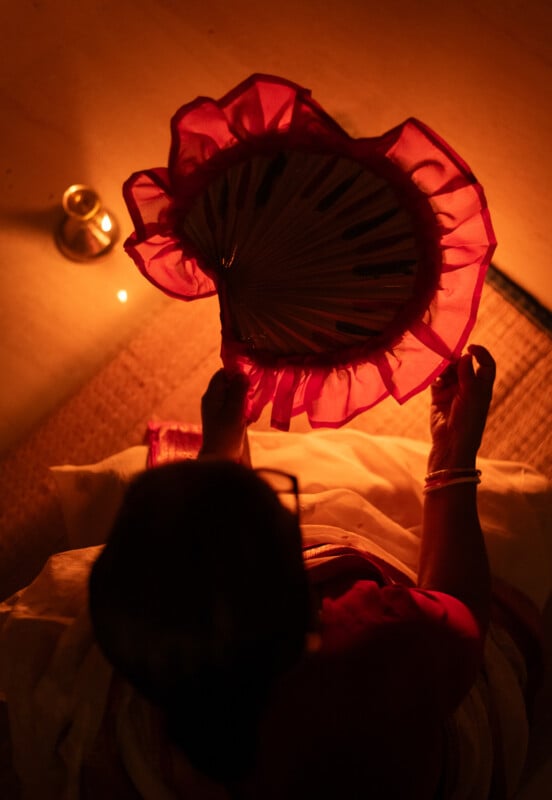 A person in traditional attire holds a decorative hand fan with red edges, illuminated by warm, soft candlelight beside a woven mat.