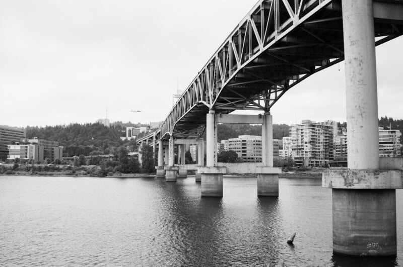 Black and white photo of a large bridge with concrete pillars spanning a river, with modern buildings and trees visible on the opposite shore under a cloudy sky.