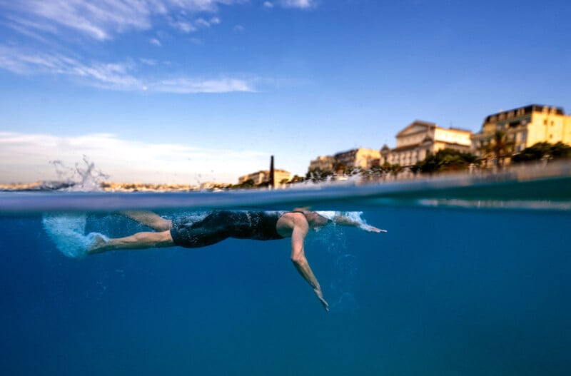 A swimmer in a black swimsuit swims freestyle near the surface of clear blue water, with a cityscape and blue sky visible in the background above the waterline.