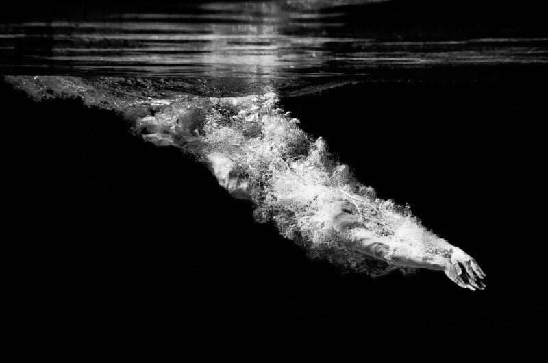 A swimmer captured underwater in black and white, arms outstretched and surrounded by a cloud of bubbles, moving gracefully through dark water beneath a rippling surface.