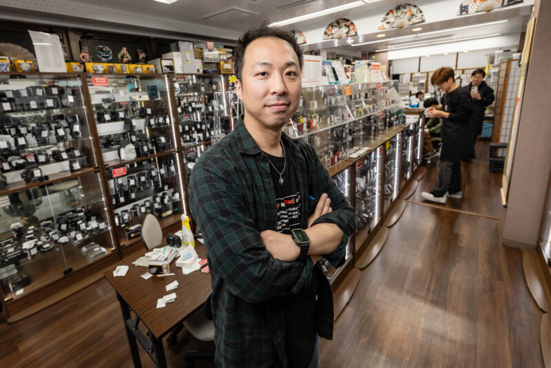 A man stands with arms crossed in a camera shop, surrounded by camera equipment and accessories. Two other people are browsing items in the background, and the shop has glass display cases and wooden floors.