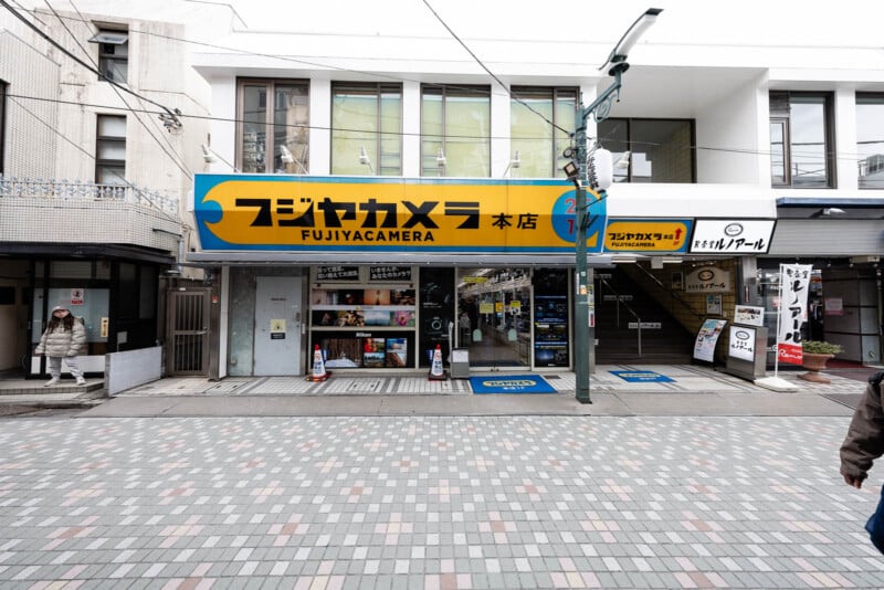Street view of Fujiyacamera store in Japan, with yellow and blue signage, glass doors, and window displays; people walk along a tiled sidewalk in front of the building.