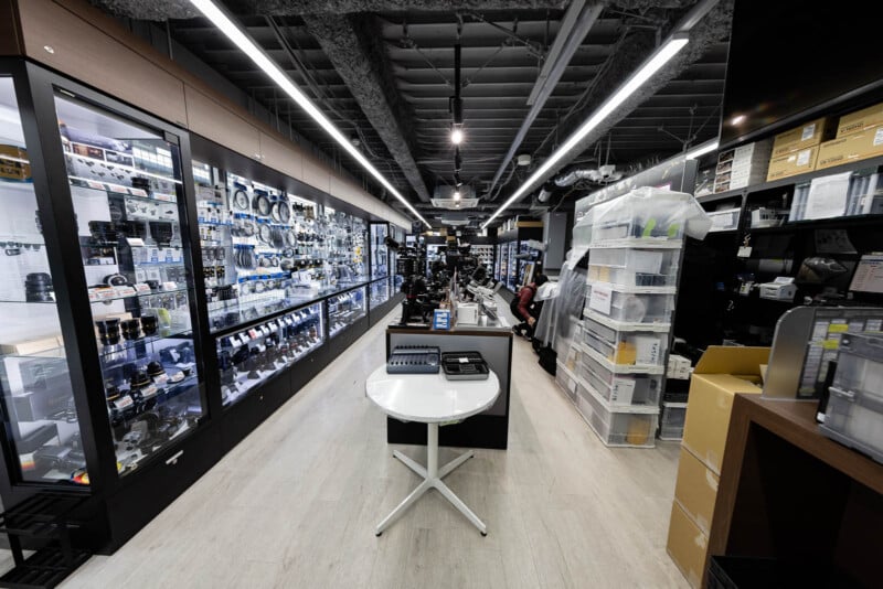 A well-lit camera store interior with glass display cases of cameras and lenses on the left, shelves with boxes on the right, and a round table with camera accessories in the center aisle.