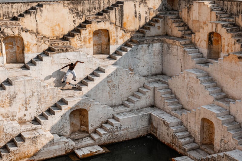 A skateboarder rides along the steep steps of an ancient stepwell with geometric patterns and weathered stone walls, casting dramatic shadows in the sunlight.