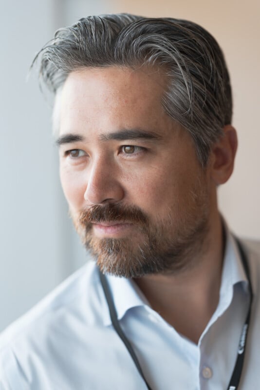 A man with gray streaked hair and a beard, wearing a light blue collared shirt and a lanyard, looks pensively out of a window with soft natural light on his face.