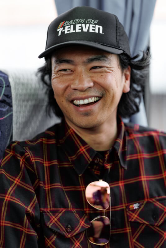 A man wearing a black "Cars of 7-Eleven" cap and a red plaid shirt smiles while sitting indoors; sunglasses hang from his shirt.